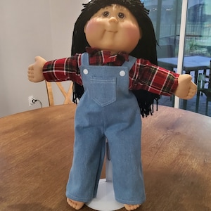 May include: A doll with dark yarn hair, brown eyes, and rosy cheeks. The doll wears a red and black plaid long-sleeve shirt under light blue denim overalls with white buttons. The doll is supported by a white metal stand and has outstretched arms.