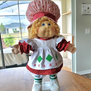 May include: A doll with blonde yarn hair and blue eyes wearing a pink strawberry-patterned bonnet, a white apron with green diamond shapes and red cross-stitch details over a red polka-dot dress, green and white striped leggings, and white shoes.