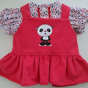 Könnte beinhalten: Rotes Babykleid mit einem Panda-Bären-Motiv auf der Vorderseite. Das Kleid hat einen gerüschten Rock und ein Oberteil mit weißen und marineblauen Sternenmustern und kurzen Ärmeln. Das Kleid hat einen runden Kragen und zwei Knöpfe.