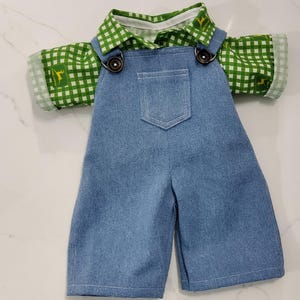 Puede incluir: Un conjunto de muñeca que presenta un peto vaquero azul con un bolsillo y botones marrones. El conjunto incluye una camisa a cuadros verdes y blancos con cuello y puños blancos. La camisa tiene un pequeño logotipo amarillo en la manga.
