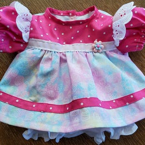 May include: A baby dress in pink and pastel shades, featuring a floral design. The dress has a pink bodice with white polka dots, short sleeves with white lace trim, and a skirt with an Easter egg pattern. A pink ribbon and flower adorn the waist.