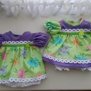 Può includere: Due completi per bambole in tessuto verde chiaro con motivo floreale rosa, blu e arancione, abbinati a maniche e dettagli del corpetto viola a pois. Ogni capo presenta bordi in pizzo bianco, un nastro verde e un piccolo bottone a fiore.