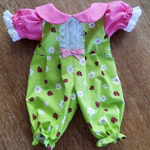 Puede incluir: Un pelele de muñeca en tela verde claro con un estampado de margaritas blancas y mariquitas rojas. Las mangas y el cuello son rosas con lunares blancos. El cuello está adornado con encaje blanco y un lazo rosa adorna el frente.