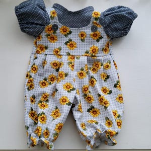 Peut inclure: Un ensemble composé d'un haut bleu foncé à pois blancs avec des manches courtes bouffantes, associé à une salopette à carreaux vichy blancs et gris avec un motif de tournesols jaunes, deux boutons foncés aux bretelles et des bas de jambes froncés.
