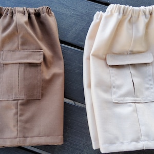 May include: Two pairs of shorts in the image. One pair is a medium brown color, and the other is a light beige. Each pair has an elastic waistband and a single square pocket on the front. The shorts are made of a smooth fabric.