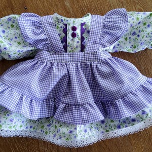 May include: A doll's dress with a purple and white gingham skirt and ruffled layers. The bodice has a floral print with purple and green details, and the sleeves are puffed. The dress is trimmed with white lace and purple buttons.
