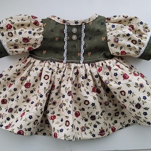 May include: A dress featuring a cream-colored fabric with a floral pattern of red and dark blue flowers. The bodice is dark olive green with a small floral print, accented by two buttons and white lace trim. It has short puff sleeves with green cuffs.