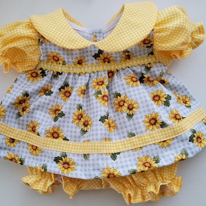 May include: A dress and bloomer set featuring a light blue and white gingham pattern with a yellow sunflower print. The dress has a rounded collar, puffed sleeves, and yellow gingham trim. The matching bloomers have a ruffled hemline.