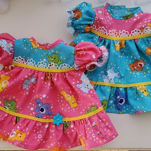 May include: Two doll dresses, one pink and one turquoise, featuring a Care Bear pattern. The dresses have frilled skirts, yellow trim, and lace details. Each dress has a small flower embellishment and puffed sleeves.