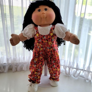 Könnte beinhalten: Eine Puppe mit schwarzem Wollhaar, braunen Augen und rosigen Wangen, bekleidet mit einem weißen kurzärmeligen Oberteil unter einer roten Latzhose mit Blumenmuster, gelben Blütenakzenten und weißen Schuhen auf einem weißen Kunststoffständer.