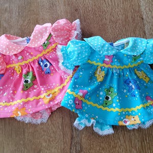 Puede incluir: Dos vestidos de muñeca, uno rosa y otro azul, con estampados de Ositos Cariñosos. El vestido rosa tiene cuello y mangas de lunares blancos, con ribete de encaje blanco. El vestido azul tiene cuello y mangas de lunares blancos, con ribete de encaje blanco.