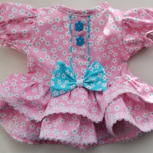 Puede incluir: Vestido rosa con estampado floral, lazo turquesa y detalles florales a juego. El vestido presenta una falda con volantes con ribete de encaje blanco y mangas cortas con volantes. Un atuendo encantador para una mascota.