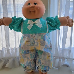 May include: A vintage doll with a round face and open arms, wearing a teal top with white polka dots and a white bow. The doll is also wearing patterned trousers with animal and letter designs.
