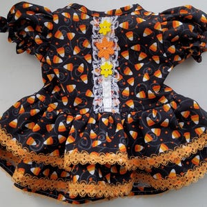 Può includere: Un vestito nero con un motivo di caramelle a forma di chicco di mais nei colori arancione, giallo e bianco. Il vestito presenta maniche corte a sbuffo, un bordo in pizzo bianco al centro e tre bottoni a forma di fiore. L'orlo ha due strati di pizzo arancione.