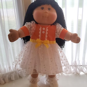 May include: A doll with black yarn hair, brown eyes, and a round face. The doll wears an orange short-sleeved bodice with two white buttons and a white lace trim. A yellow ribbon bow sits at the waist above a white skirt with a floral pattern.