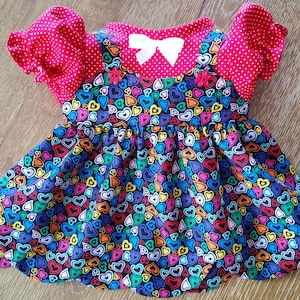 May include: A vibrant doll dress with a heart design in multiple colours on a dark blue base. The top is red with white polka dots and ruffled sleeves. A white bow and two small flower accents adorn the neckline.