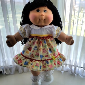 Puede incluir: Una muñeca de piel oscura, cabello de lana negra y mejillas sonrosadas que viste una camisa blanca con cuello y mangas abullonadas, y un vestido amarillo con un patrón colorido de pequeñas prendas, acabado con un borde rojo y una pequeña flor amarilla.