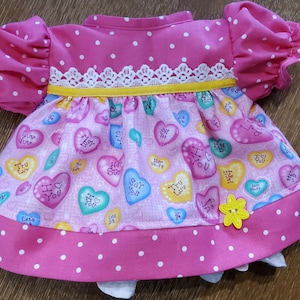 May include: A pink doll dress with white polka dots and a heart-themed pattern. The dress features puffed sleeves, a white lace trim, and a yellow ribbon. The skirt is adorned with colorful heart-shaped candies with phrases like "I Love You". A yellow flower is sewn on the skirt.