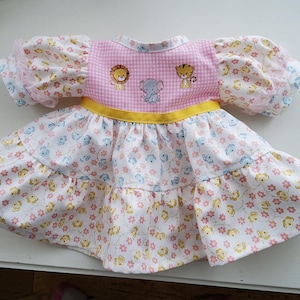 May include: A dress featuring a pink and white gingham bodice with embroidered lion, elephant, and tiger motifs. It has a yellow waistband, puffed sleeves, and a tiered skirt patterned with small pink flowers and yellow shapes on a white background.