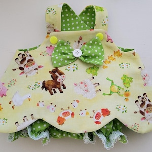 Könnte beinhalten: Ein gelb-grünes Lätzchen mit Bauernhoftieren, mit einer grünen Schleife und Verzierungen mit Polka Dots. Das Lätzchen zeigt Illustrationen von Kühen, Schafen, Schweinen, Pferden und Fröschen. Das Lätzchen hat einen gewellten Rand mit weißer Spitzentrimmung.