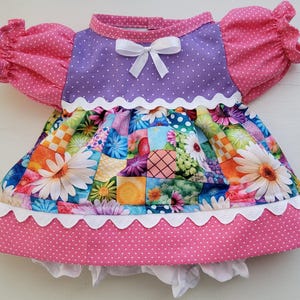 Könnte beinhalten: Ein Puppenkleid mit einem violetten, gepunkteten Oberteil mit weißer Schleife, rosa gepunkteten Puffärmeln und einem Rock mit buntem Blumen-Patchwork-Muster. Das Kleid ist mit weißer Bogenkante verziert und enthält eine weiße Pumphose.