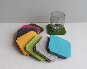 May include: A set of eight felt coasters in various colors, including green, gray, yellow, turquoise, pink, and black. The coasters are square with rounded corners and are arranged in a fan shape. A clear glass is sitting on a green coaster.