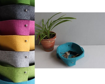 May include: A stack of felt storage baskets in various colors, including green, gray, pink, yellow, and teal. Each basket has a silver button closure. A teal basket with keys inside is on a table next to a potted plant.