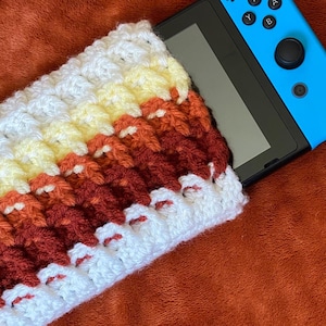 Könnte beinhalten: Eine gehäkelte Hülle in einem Farbverlauf aus Weiß, Gelb, Orange und Burgunder, die teilweise ein blaues Handheld-Spielgerät bedeckt. Die Hülle hat ein strukturiertes, geripptes Muster.