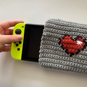 Puede incluir: Una funda de crochet gris con un diseño de corazón pixelado rojo. La funda cubre parcialmente una consola Nintendo Switch amarilla.