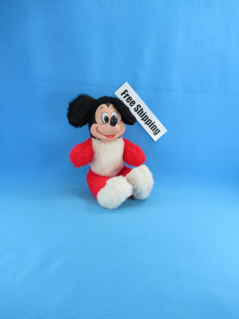 Peut inclure: Une peluche Mickey Mouse assis, avec un corps rouge, des d&eacute;tails blancs et des oreilles noires. Le jouet a un visage souriant et est pos&eacute; sur un fond bleu. Une &eacute;tiquette blanche avec les mots "Free Shipping" est &eacute;galement pr&eacute;sente.