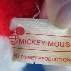 Peut inclure: Gros plan sur une &eacute;tiquette d'une peluche Mickey Mouse. L'&eacute;tiquette est de couleur cr&egrave;me avec le texte rouge "MICKEY MOUSE" et "WALT DISNEY PRODUCTIONS". La peluche a des poils rouges et blancs.