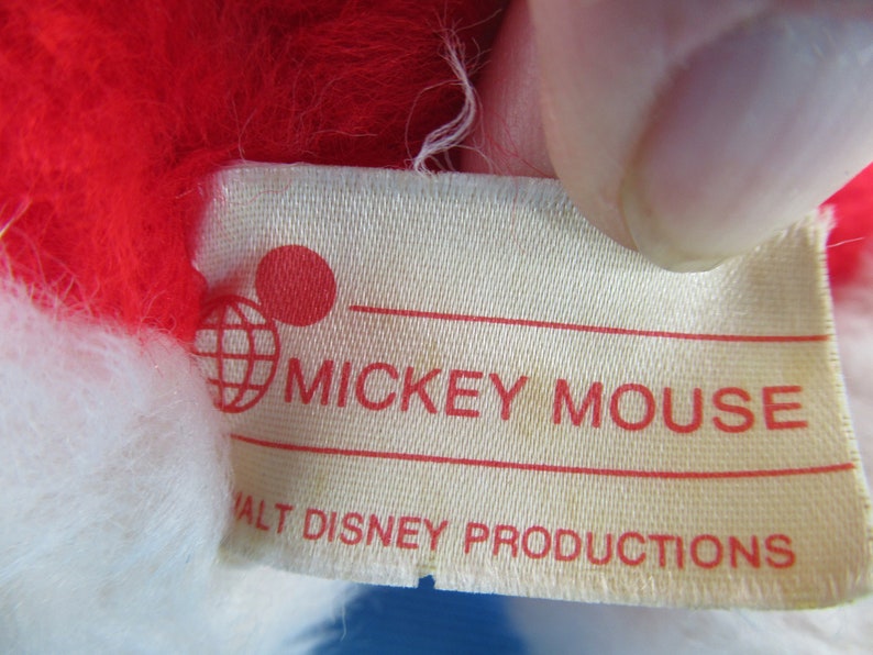 Peut inclure: Gros plan sur une &eacute;tiquette d'une peluche Mickey Mouse. L'&eacute;tiquette est de couleur cr&egrave;me avec le texte rouge "MICKEY MOUSE" et "WALT DISNEY PRODUCTIONS". La peluche a des poils rouges et blancs.
