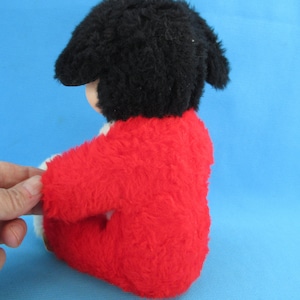 Peut inclure: Une poup&eacute;e en peluche vintage avec un bonnet noir et un manteau rouge vif. La poup&eacute;e a un teint clair et est assise. Le manteau de la poup&eacute;e a une bordure blanche. Le fond est bleu clair uni.
