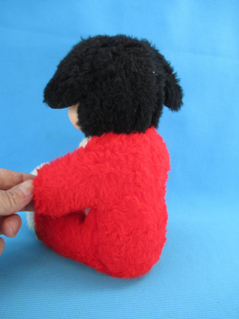 Peut inclure: Une poup&eacute;e en peluche vintage avec un bonnet noir et un manteau rouge vif. La poup&eacute;e a un teint clair et est assise. Le manteau de la poup&eacute;e a une bordure blanche. Le fond est bleu clair uni.
