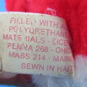 Peut inclure: Gros plan d'une &eacute;tiquette avec du texte rouge sur fond cr&egrave;me. L'&eacute;tiquette indique "FILLED WITH ALL POLYURETHANE MATERIALS - LICEN PENNA 268 - OHIO MASS 214 - MAINE SEWN IN HAITI". L'&eacute;tiquette est attach&eacute;e &agrave; une peluche rouge et blanche.