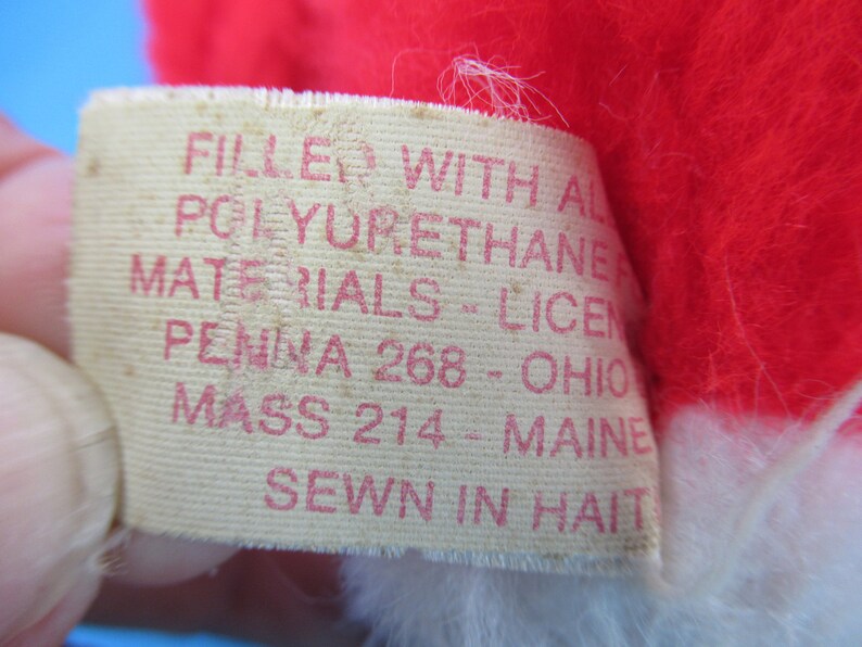 Peut inclure: Gros plan d'une &eacute;tiquette avec du texte rouge sur fond cr&egrave;me. L'&eacute;tiquette indique "FILLED WITH ALL POLYURETHANE MATERIALS - LICEN PENNA 268 - OHIO MASS 214 - MAINE SEWN IN HAITI". L'&eacute;tiquette est attach&eacute;e &agrave; une peluche rouge et blanche.