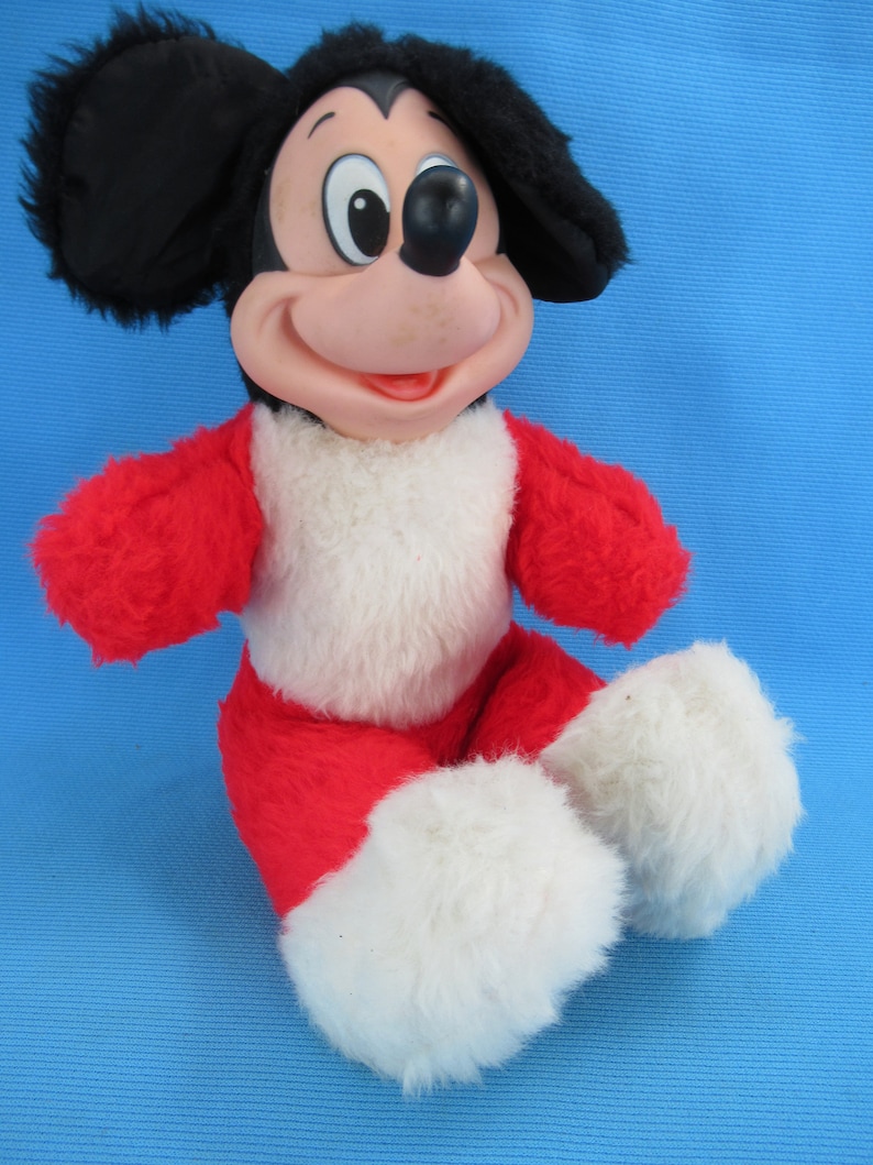 Peut inclure: Une peluche vintage de Mickey Mouse. La peluche pr&eacute;sente une t&ecirc;te noire avec de grandes oreilles, un visage rose et une tenue rouge et blanche. Le jouet est assis, les jambes tendues.