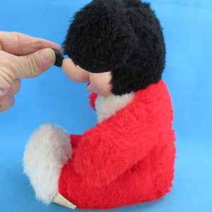 Peut inclure: Un jouet en peluche vintage avec une t&ecirc;te noire et poilue, un corps rouge et des accents blancs. Le jouet a un nez rond et noir et une &eacute;tiquette attach&eacute;e en bas. Le jouet est pos&eacute; sur une surface bleue.