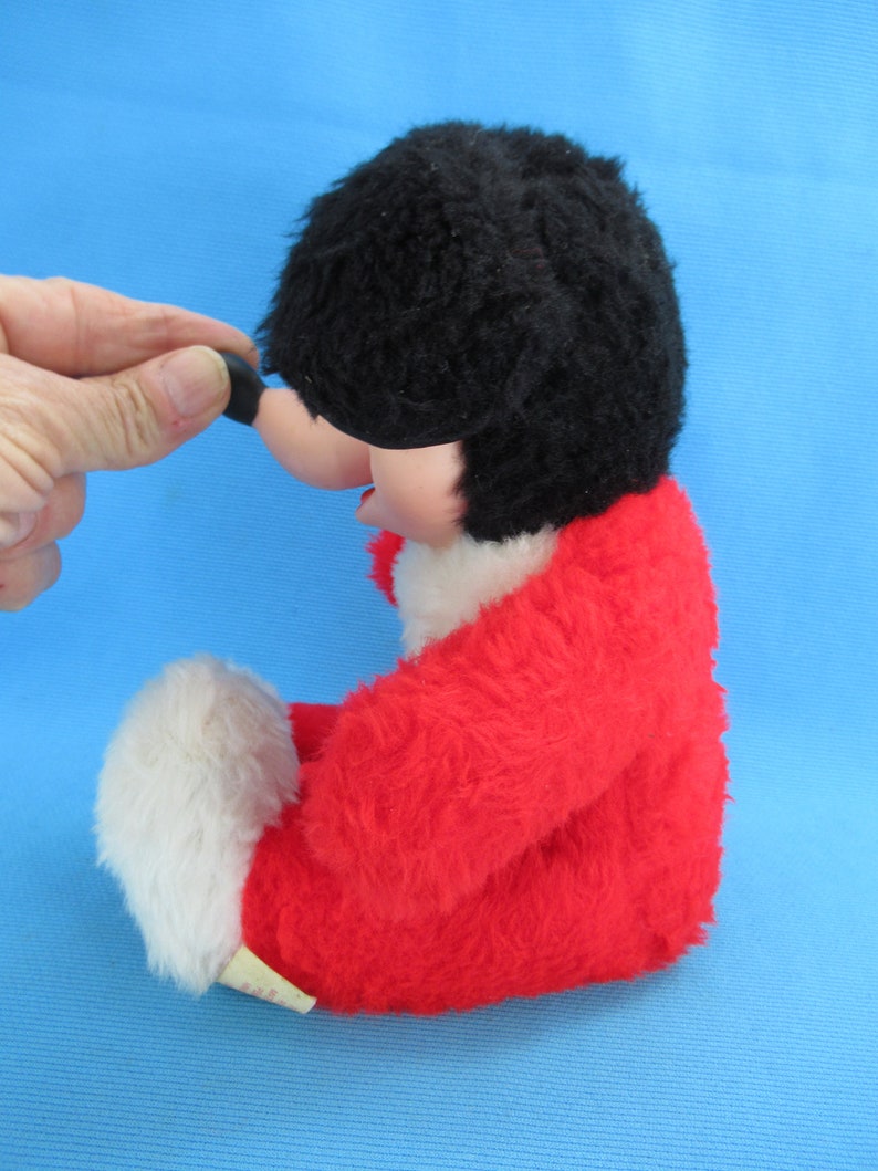 Peut inclure: Un jouet en peluche vintage avec une t&ecirc;te noire et poilue, un corps rouge et des accents blancs. Le jouet a un nez rond et noir et une &eacute;tiquette attach&eacute;e en bas. Le jouet est pos&eacute; sur une surface bleue.