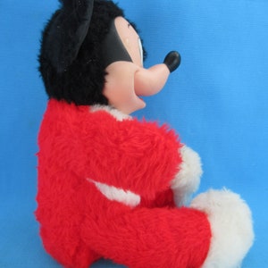 Peut inclure: Une peluche vintage de Mickey Mouse, avec un corps rouge, des pieds blancs et une t&ecirc;te noire avec de grandes oreilles. Le jouet a une expression souriante et est pos&eacute; sur un fond bleu.