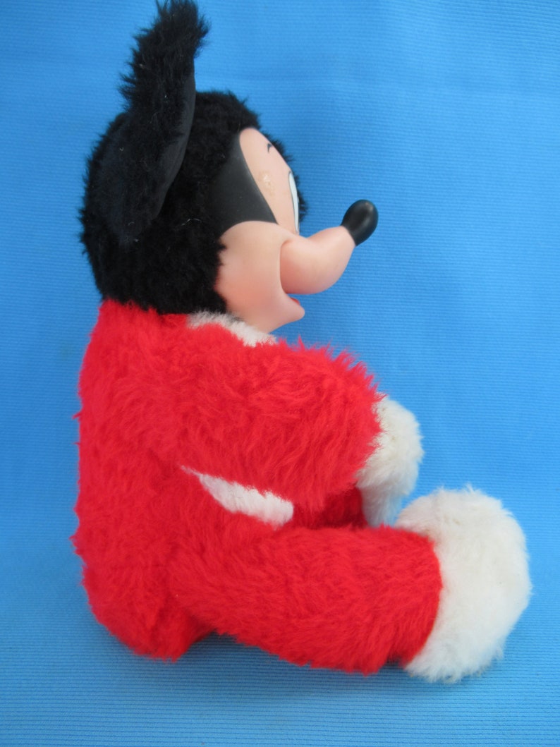 Peut inclure: Une peluche vintage de Mickey Mouse, avec un corps rouge, des pieds blancs et une t&ecirc;te noire avec de grandes oreilles. Le jouet a une expression souriante et est pos&eacute; sur un fond bleu.