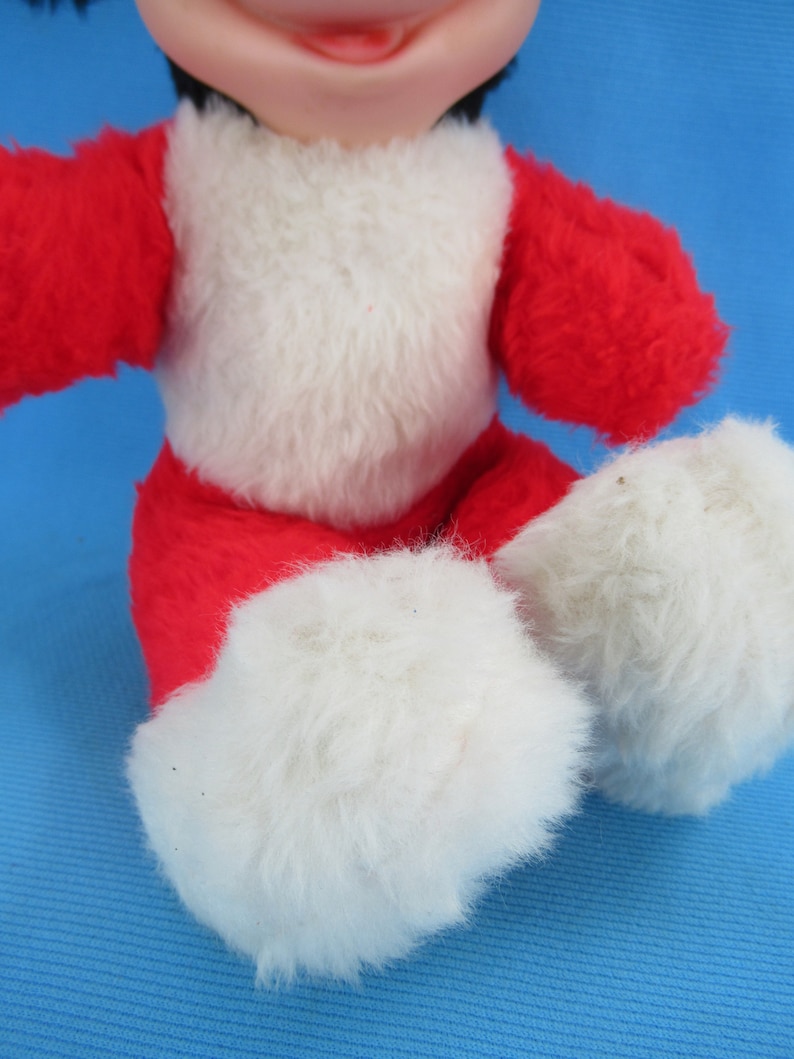 Peut inclure: Une poup&eacute;e en peluche vintage avec une tenue rouge et blanche. La poup&eacute;e a des cheveux noirs et un visage souriant. Le corps de la poup&eacute;e est rouge avec une poitrine blanche et des pieds blancs et duveteux. La poup&eacute;e est assise.