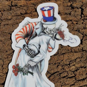 May include: A sticker featuring a skeleton couple dressed in a wedding gown and a red, white, and blue suit with a top hat. The skeleton man is holding a bouquet of red roses and the skeleton woman is holding a single red rose. The sticker is on a wooden background.