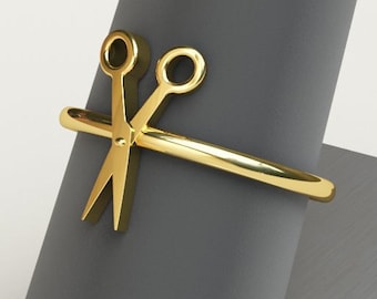 May include: A gold-colored ring with a pair of scissors design. The ring is on a gray display stand.