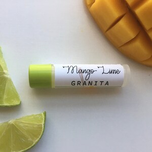 Puede incluir: Un tubo de bálsamo labial verde con una etiqueta blanca que dice "Mango-Lime Granita". En el fondo hay dos rodajas de lima y un trozo de mango cortado en dados.