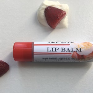 May include: A red and white lip balm tube with the text "Shortcake Lip Balm" and a graphic of a strawberry shortcake. The ingredients are listed as "Organic Sunflower Oil, Organic Coconut Oil, Beeswax, Vanilla Infusion."