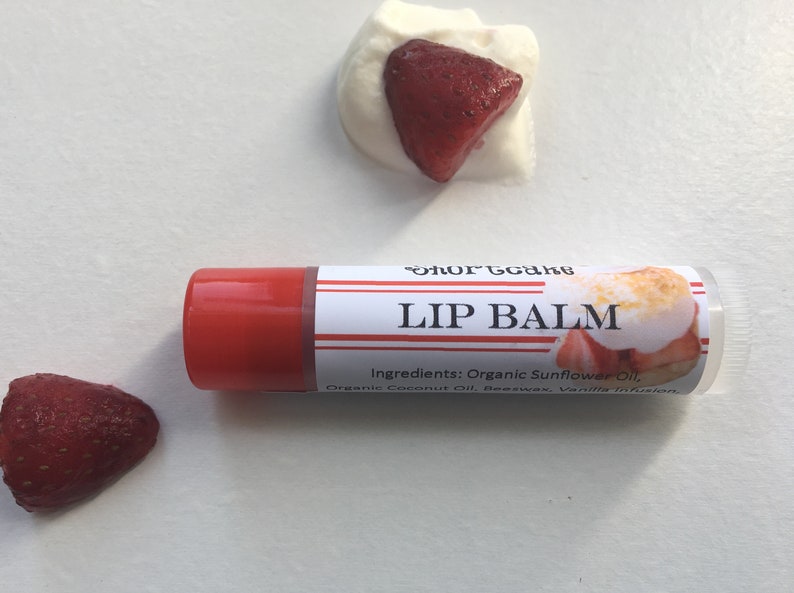 May include: A red and white lip balm tube with the text "Shortcake Lip Balm" and a graphic of a strawberry shortcake. The ingredients are listed as "Organic Sunflower Oil, Organic Coconut Oil, Beeswax, Vanilla Infusion."