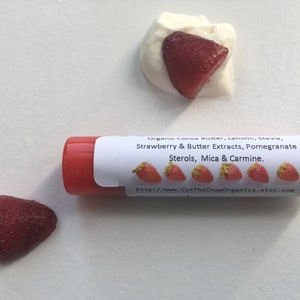 May include: A red lip balm tube with a white label that reads "Organic Cocoa Butter, Lanolin, Stevia, Strawberry & Butter Extracts, Pomegranate Sterols, Mica & Carmine." The label also includes a website address: "http://wwww.CutTheCrapOrganics.etsy.com". There are five small red strawberry images on the label.