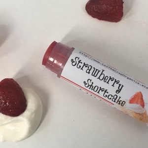 May include: A clear tube of lip balm with a red cap and a white label that says "Strawberry Shortcake" and a website address. The label also features a small illustration of a strawberry shortcake dessert.