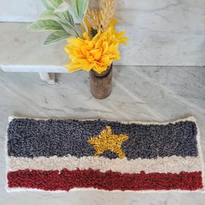 May include: Handmade rectangular rug with a star design. The rug features a blue, white, and red striped pattern, with a yellow star in the center. The rug is made of a textured material and is likely used as a decorative accent.